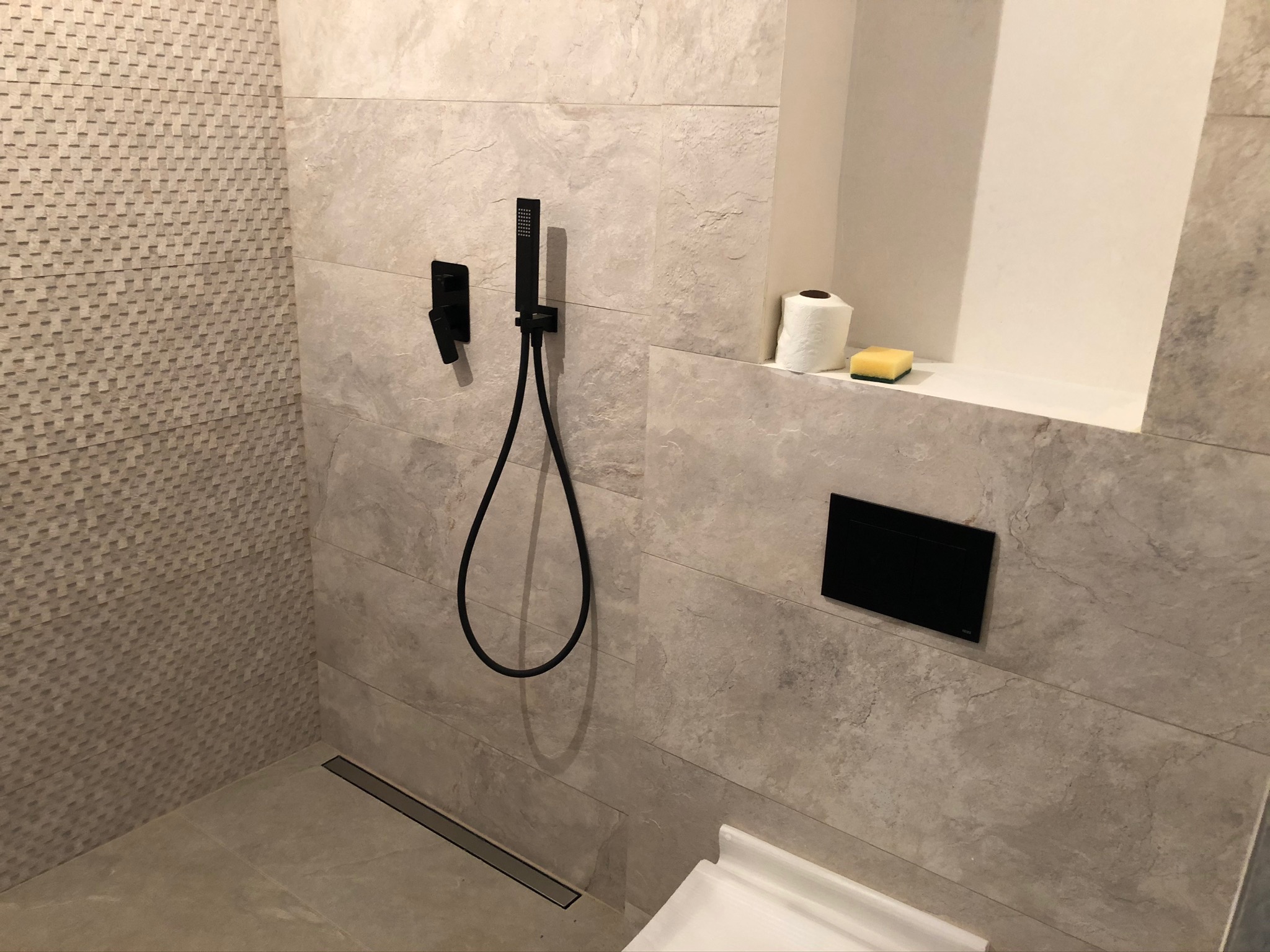 Nowoczesna kabina prysznicowa z czarną armaturą, płytki imitujące kamień, odpływ liniowy, półka z rolką papieru toaletowego i gąbką.