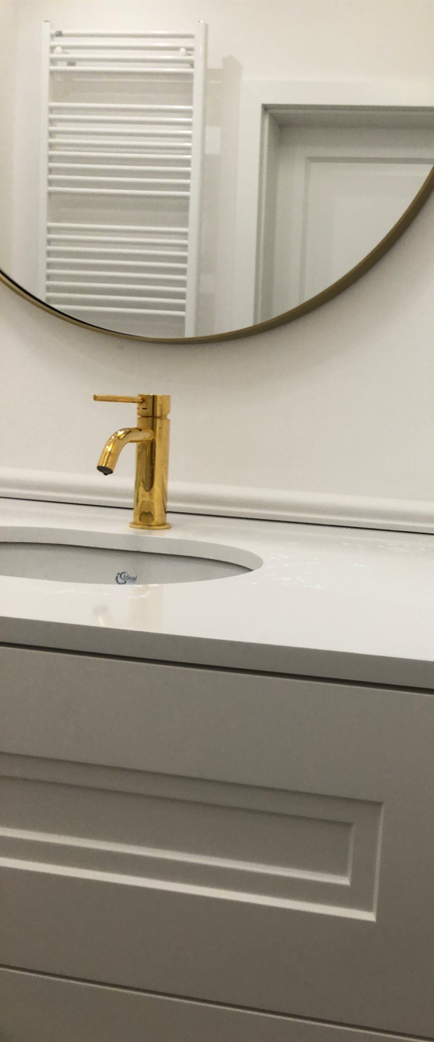 Bateria umywalkowa w kolorze złotym, umieszczona na białym blacie umywalkowym, z widocznym fragmentem okrągłego lustra i grzejnika drabinkowego w tle.