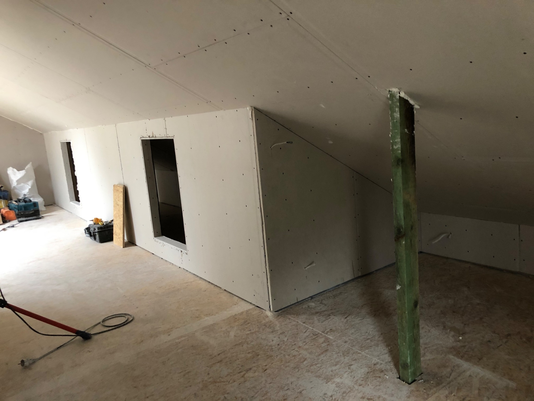 Poddasze w trakcie adaptacji: ściany i sufit wyłożone płytami gipsowo-kartonowymi, widoczne otwory okienne, drewniany słup podtrzymujący konstrukcję dachu, narzędzia i materiały budowlane w tle.