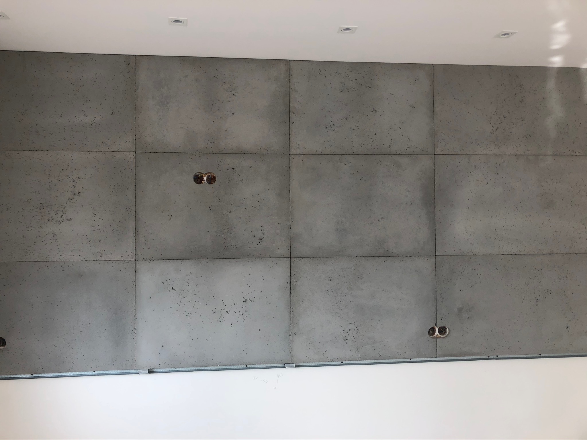 Ściana wykończona panelami imitującymi beton, widoczne puszki elektryczne bez osprzętu oraz biały sufit z oświetleniem punktowym.