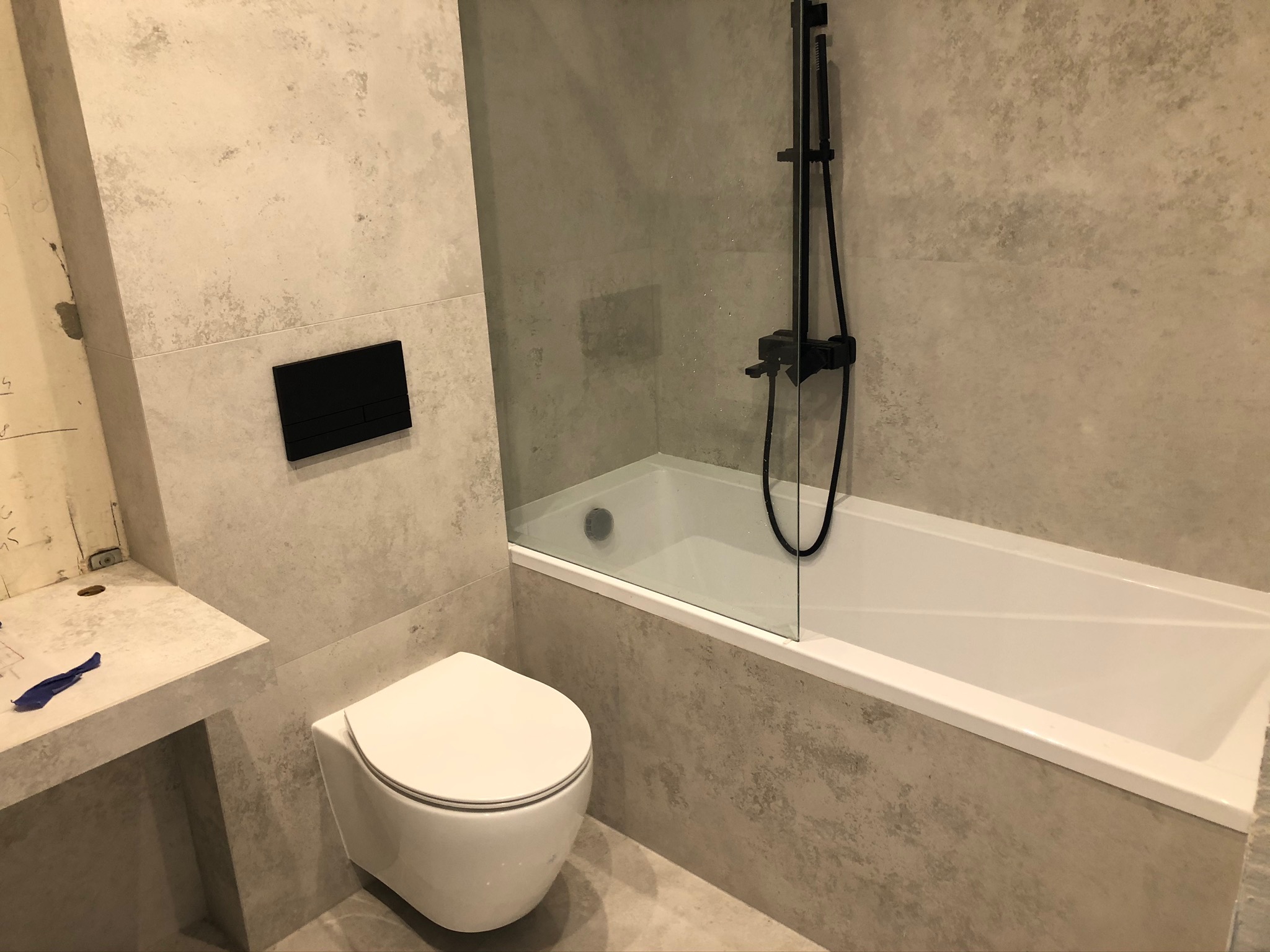 Nowoczesna łazienka z szarą, teksturowaną okładziną ścienną, wiszącą toaletą i czarną armaturą prysznicową za szklanym panelem.