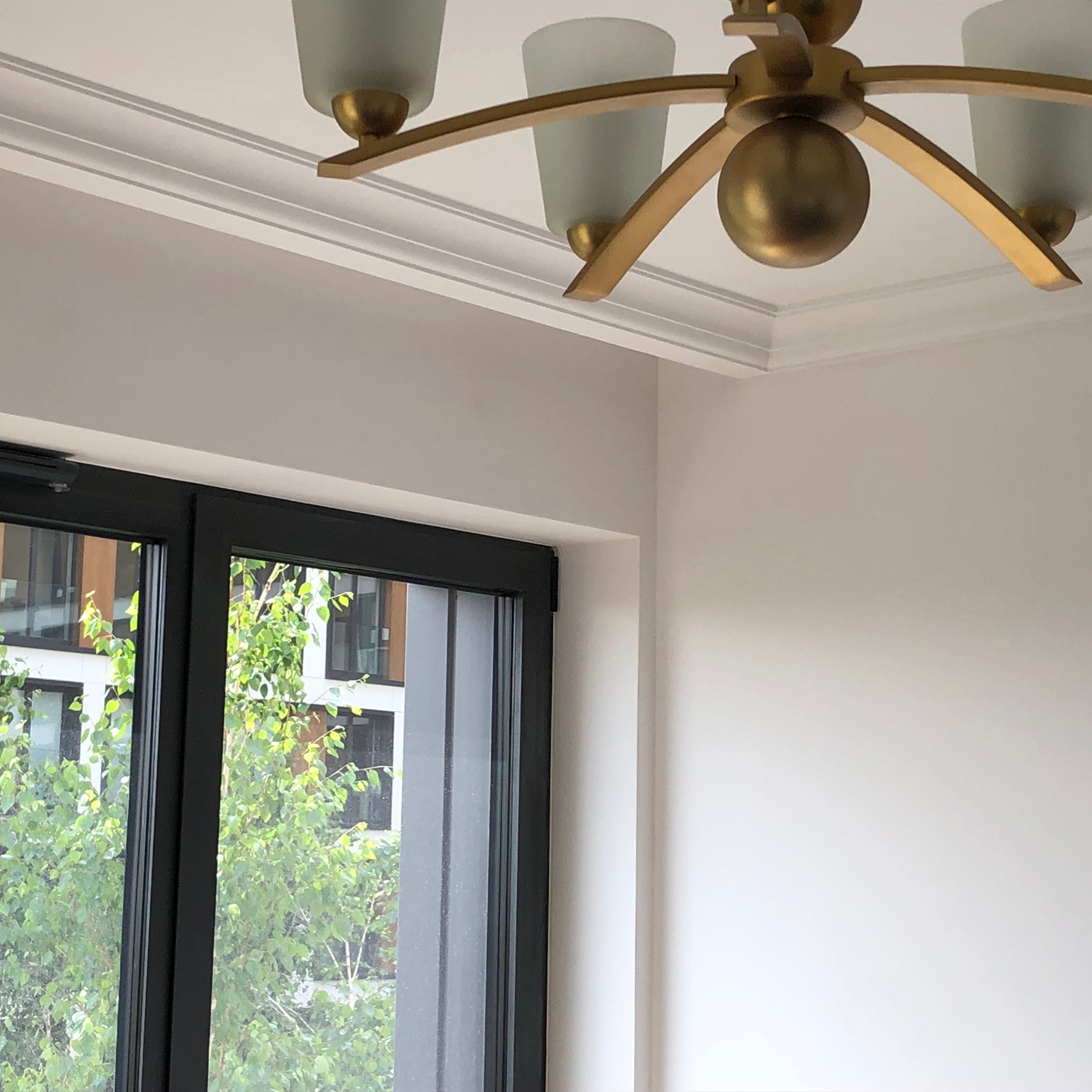 Fragment pokoju z białymi ścianami, ozdobnym gzymsem sufitowym i złotą lampą wiszącą z mlecznymi kloszami, widok na zieleń za oknem.