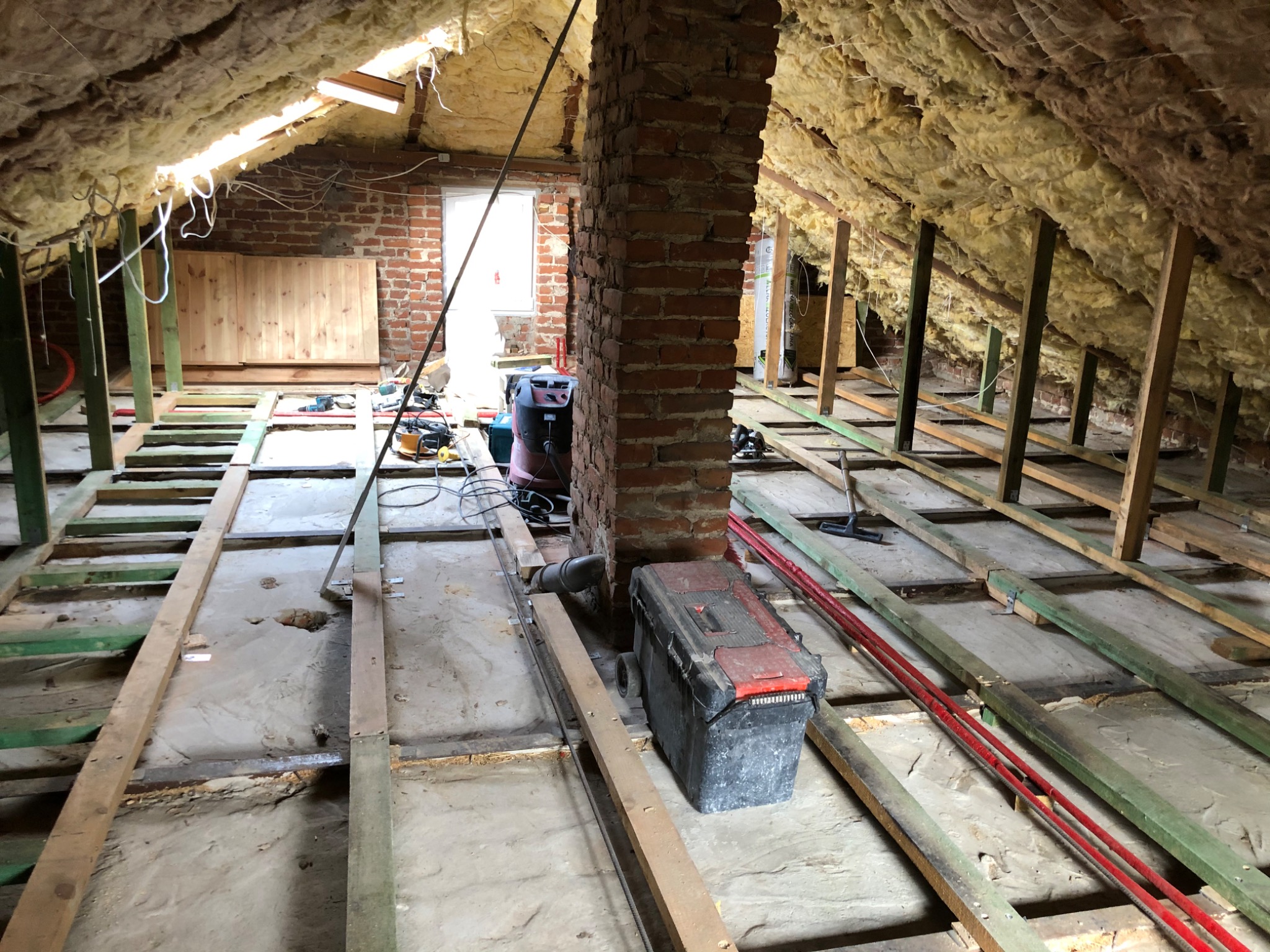 Poddasze w trakcie ocieplania: wełna mineralna izolująca skośny dach, drewniany szkielet konstrukcyjny podłogi, odsłonięty ceglany komin, narzędzia i skrzynka narzędziowa.