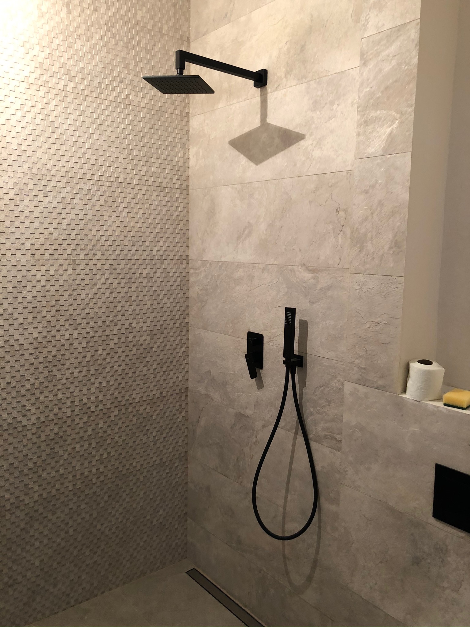 Nowoczesna kabina prysznicowa z deszczownicą w kolorze czarnym, wykończona płytkami w odcieniach beżu i szarości, z odpływem liniowym w podłodze i akcesoriami łazienkowymi.