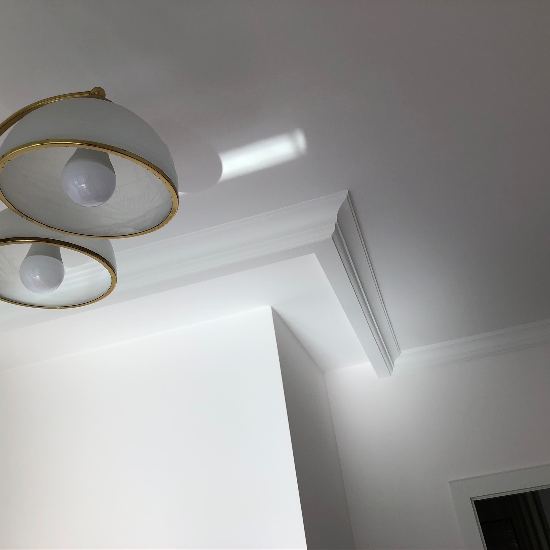 Nowoczesne oświetlenie sufitowe z dwoma kloszami z białego szkła i złotymi obwódkami, widoczne sztukaterie sufitowe i gładkie, białe ściany w jasnym wnętrzu.