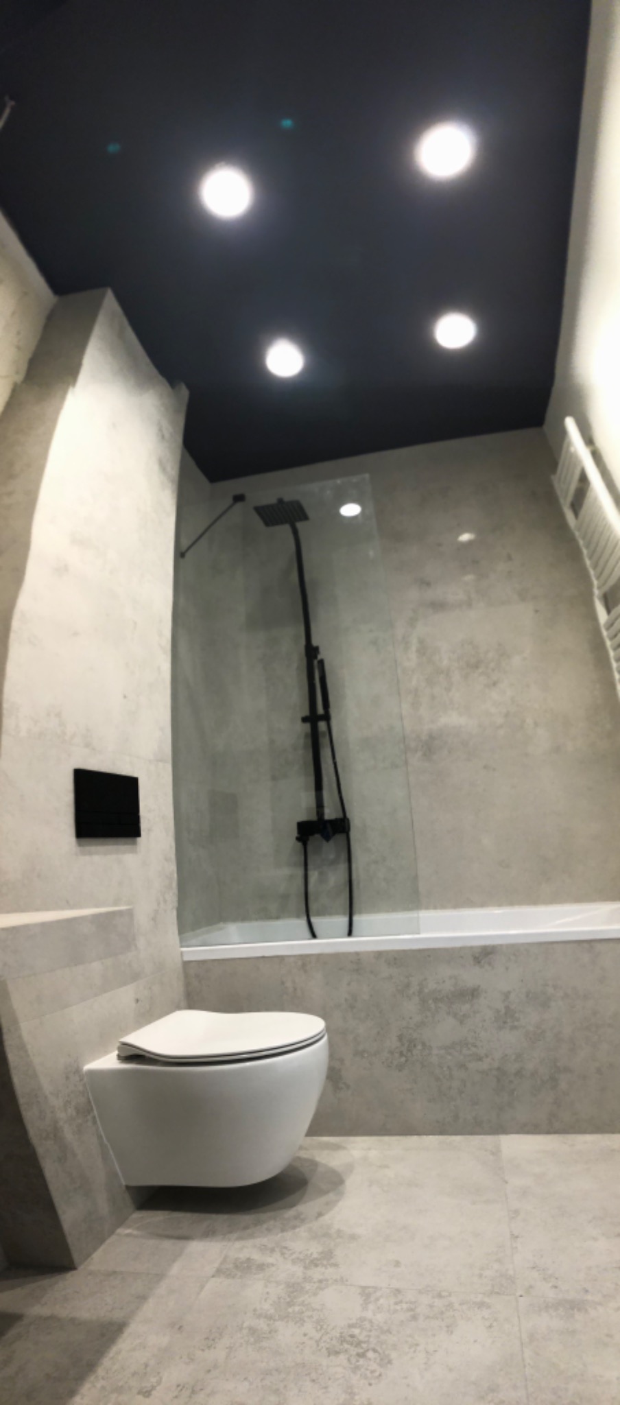 Nowoczesna łazienka z wiszącą toaletą, czarną armaturą prysznicową i szarą glazurą imitującą beton.