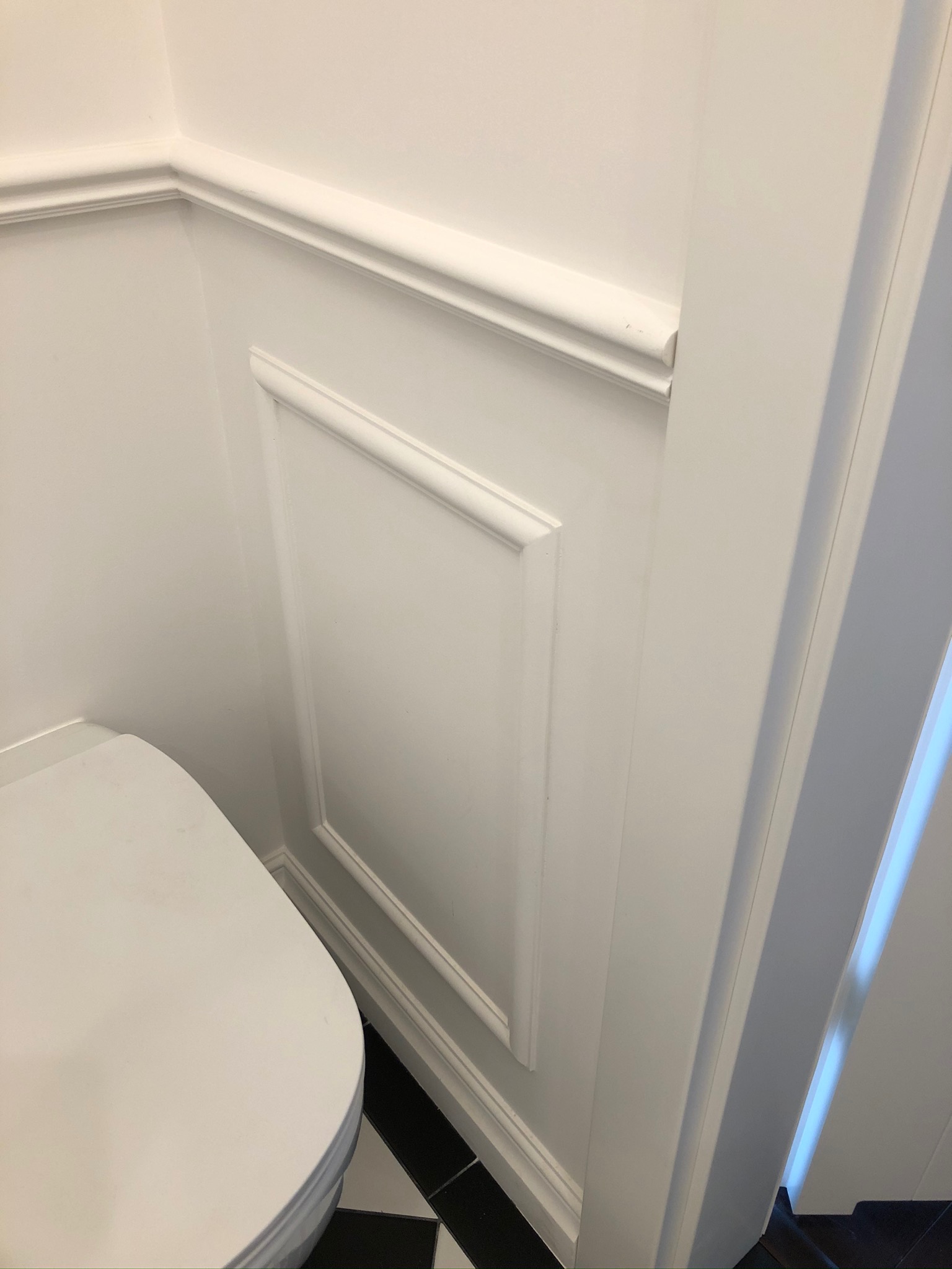 Fragment toalety z białymi ścianami ozdobionymi sztukaterią i czarno-białą podłogą w szachownicę, widoczny także fragment muszli klozetowej.