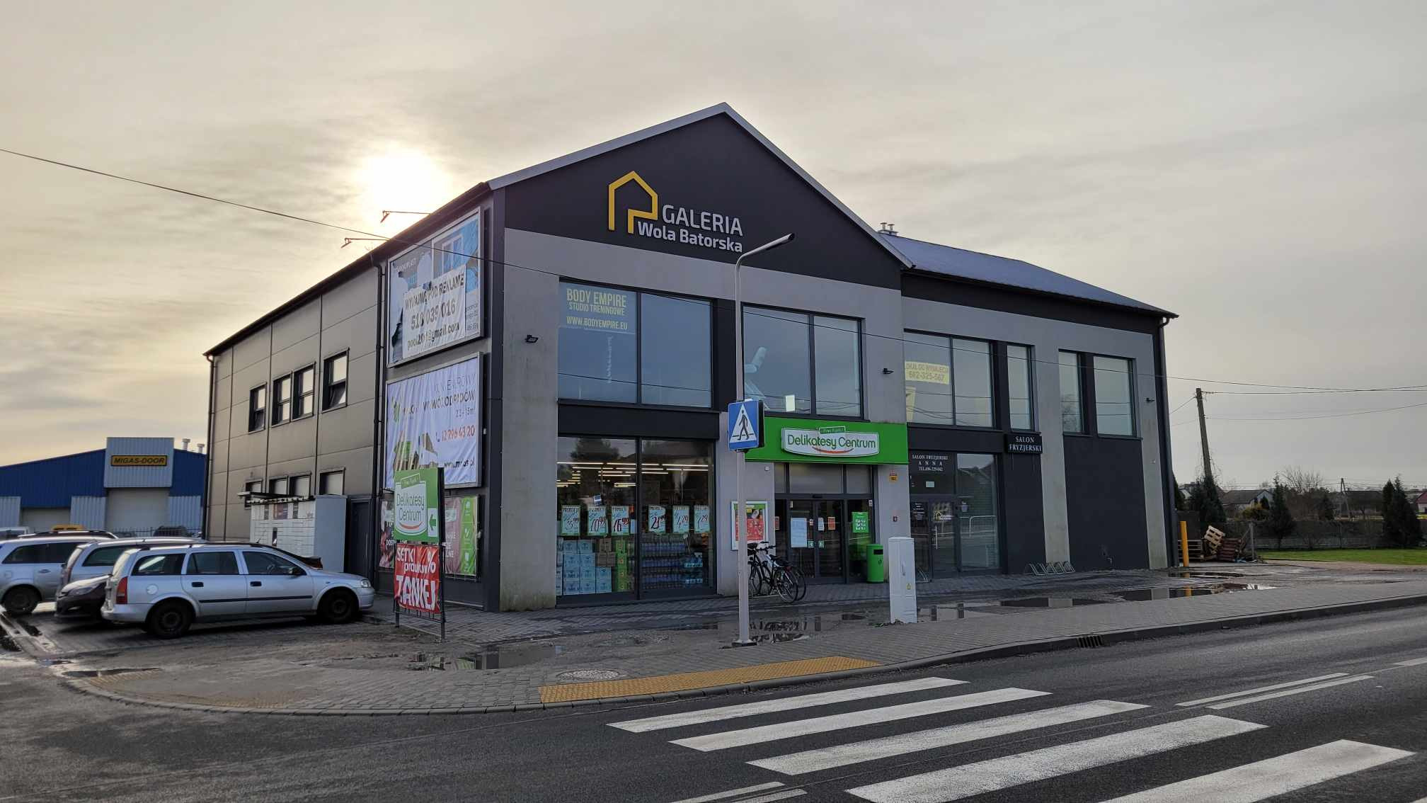 Zewnętrzny widok parterowego budynku usługowego Galeria Wola Batorska z szyldem i reklamami na elewacji, widoczny chodnik i ulica z przejściem dla pieszych.