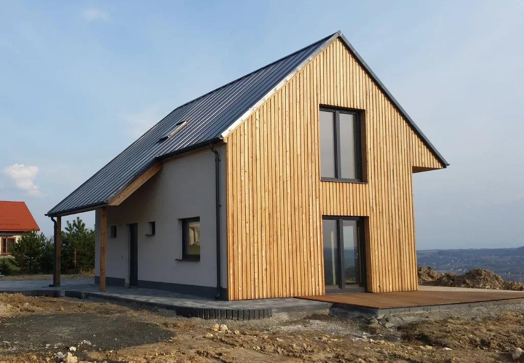 Nowoczesny dom jednorodzinny z drewnianą elewacją, ciemnym dachem i dużymi oknami, otoczony świeżo zagospodarowanym terenem.