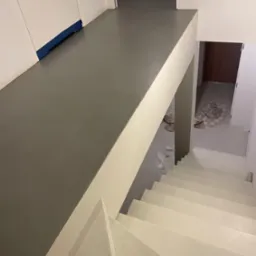 Szara, gładka posadzka na podeście schodowym i schody pomalowane na jasny kolor, widok z góry, zabezpieczenie folią przy ścianie, pozostałości po remoncie w tle.