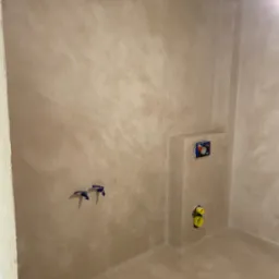 Beżowa ściana i podłoga w łazience wykończone z efektem dekoracyjnym, widoczne instalacje wodno-kanalizacyjne i niebieska taśma malarska przy suficie.