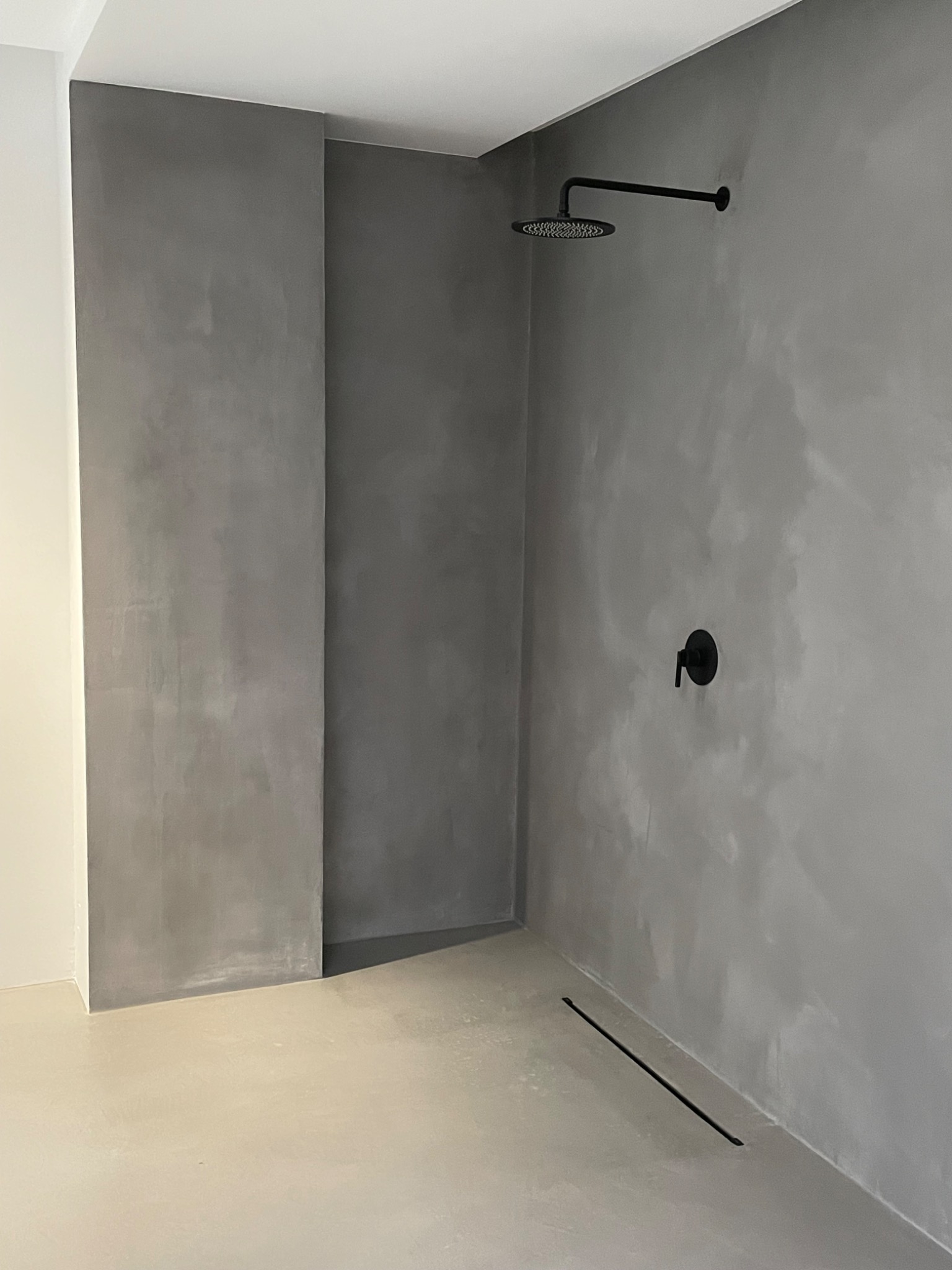 Minimalistyczna kabina prysznicowa z szarą, teksturowaną ścianą i podłogą, czarna armatura i liniowy odpływ, surowe wykończenie.