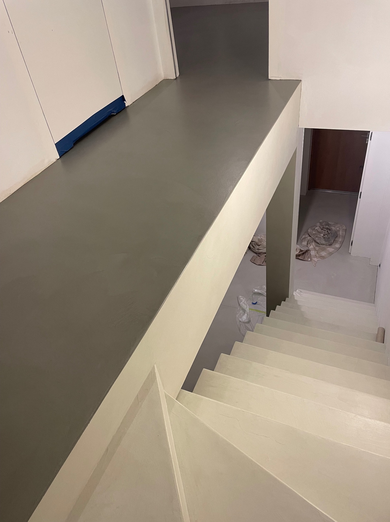 Szara, gładka posadzka na podeście schodowym i schody pomalowane na jasny kolor, widok z góry, zabezpieczenie folią przy ścianie, pozostałości po remoncie w tle.