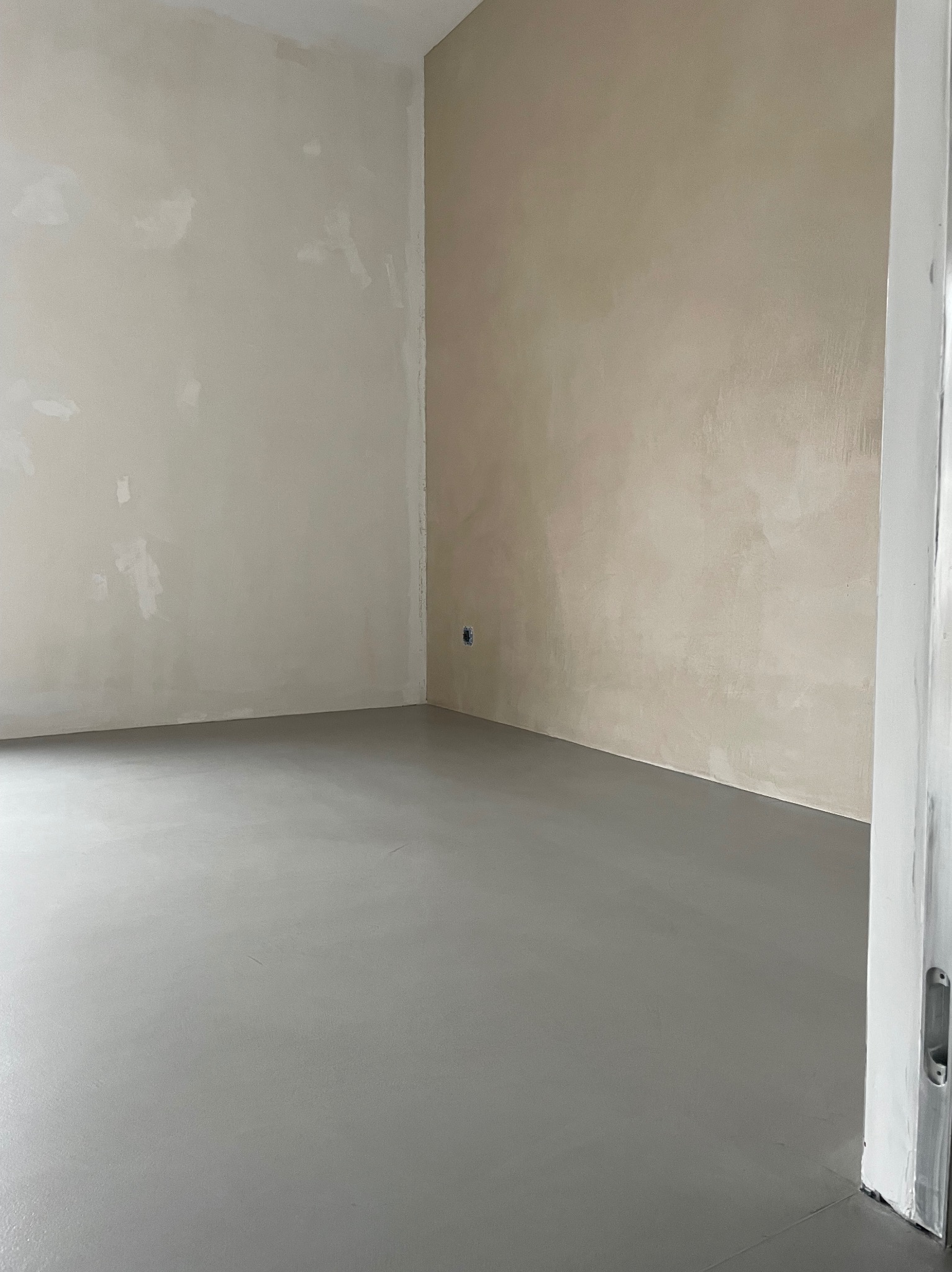 Wnętrze pokoju z szarą, gładką posadzką i ścianami w odcieniach beżu, widoczny fragment futryny drzwi.