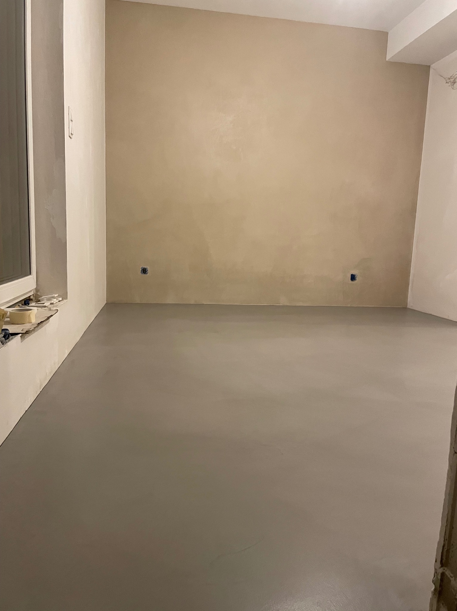 Szara, gładka posadzka w pustym pokoju z beżową ścianą i widocznymi puszkami elektrycznymi, taśma malarska na parapecie okna.