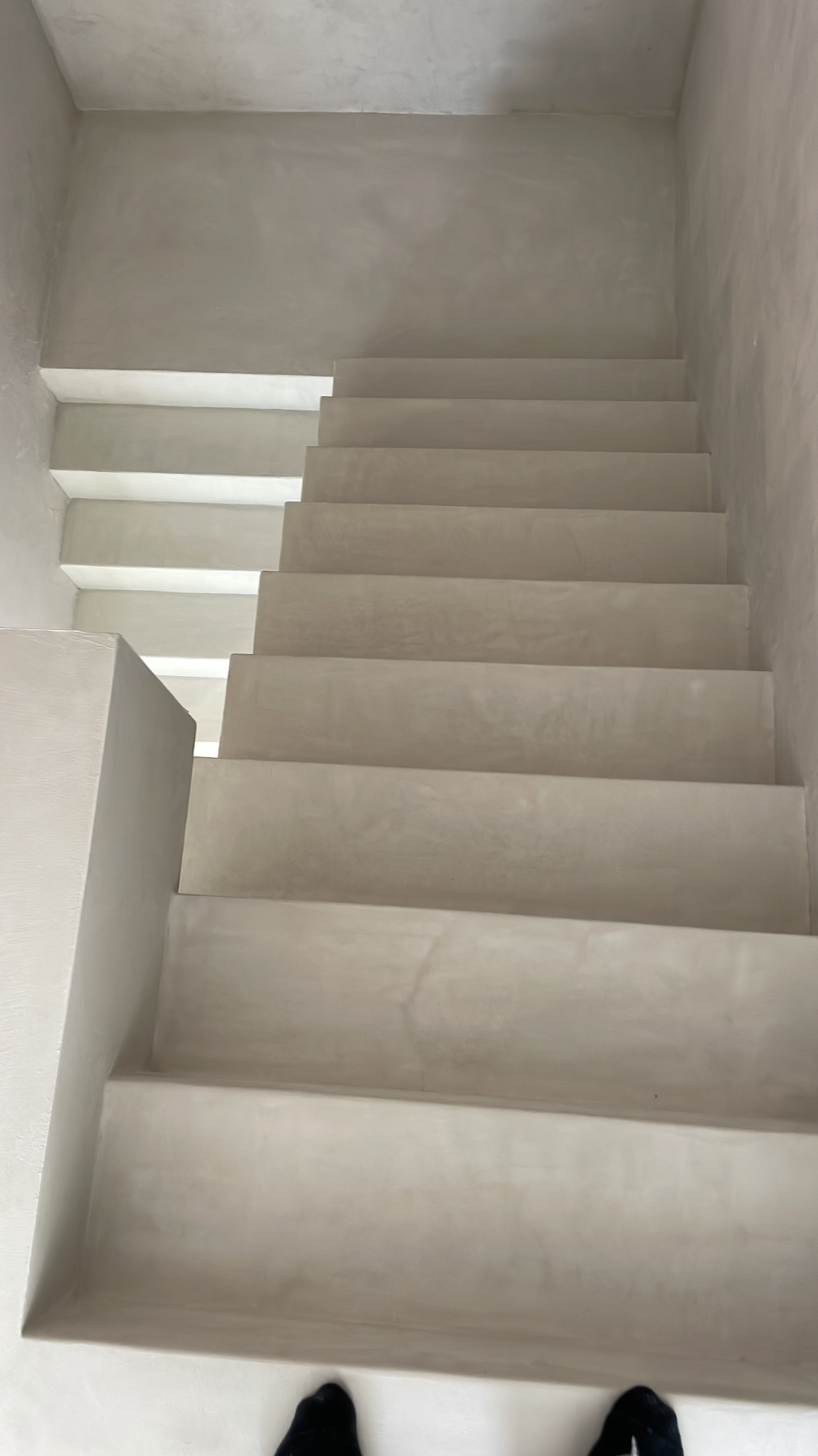 Minimalistyczne schody wykończone jasnoszarą, gładką powierzchnią, widoczne stopy w czarnych skarpetach na dole kadru.