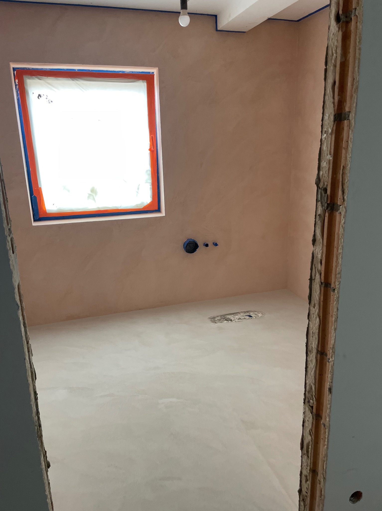 Łazienka w trakcie wykańczania: ściany i podłoga pokryte jasną, beżową warstwą, okno zabezpieczone folią malarską, widoczne wystające rury instalacji wodnej, surowa futryna drzwi.