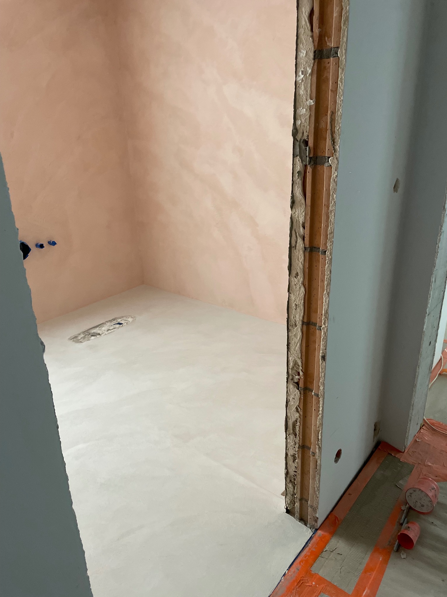 Łazienka w trakcie wykończenia mikrocementem w odcieniach różu i bieli, widoczne niedokończone obramowanie drzwi i instalacja wodna.