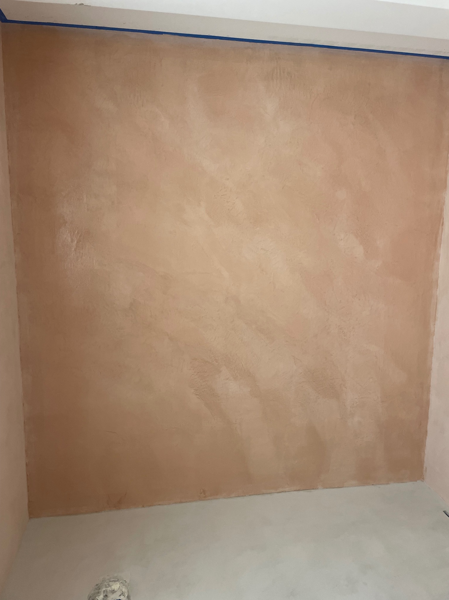 Beżowa ściana z efektem dekoracyjnym, zabezpieczona taśmą malarską przy suficie, jasna wylewka podłogowa w rogu pomieszczenia.