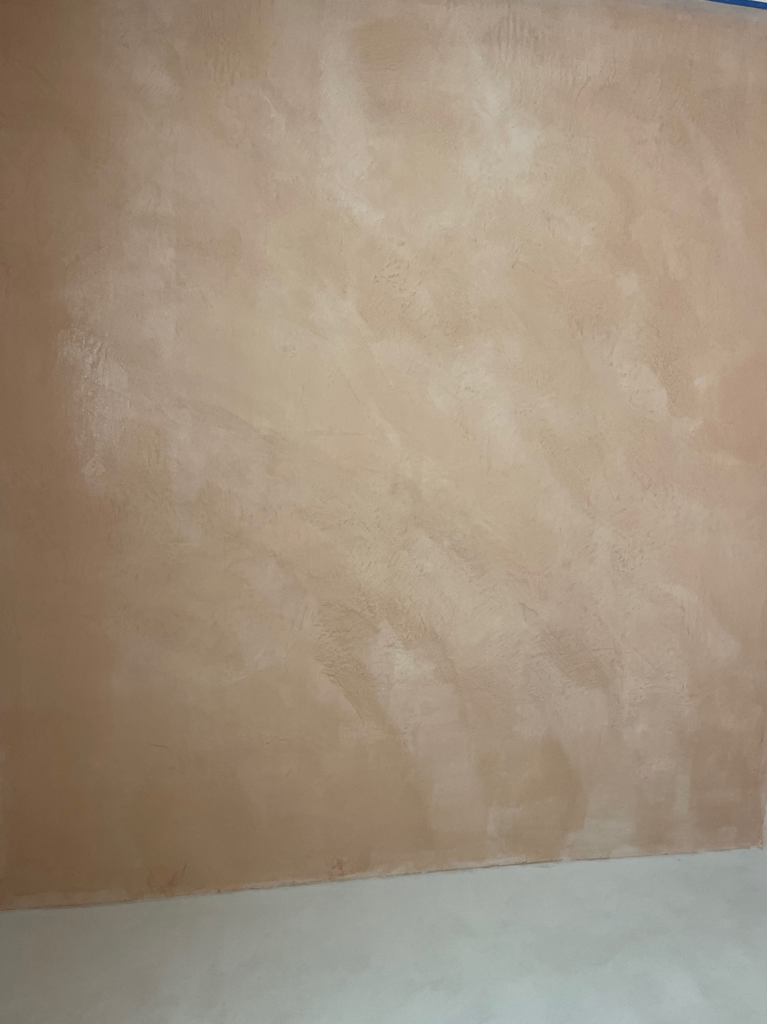 Ściana wykończona beżową, nieregularną fakturą, widoczna dolna krawędź ściany i fragment podłogi pomalowanej na jasny kolor.