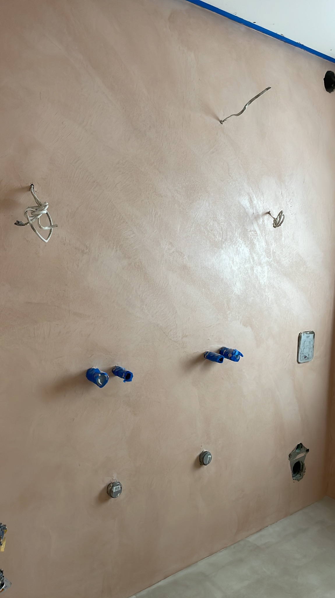 Ściana pokryta dekoracyjną, beżową powłoką z widocznymi instalacjami elektrycznymi i hydraulicznymi zabezpieczonymi niebieską taśmą, przygotowana do dalszych prac wykończeniowych.