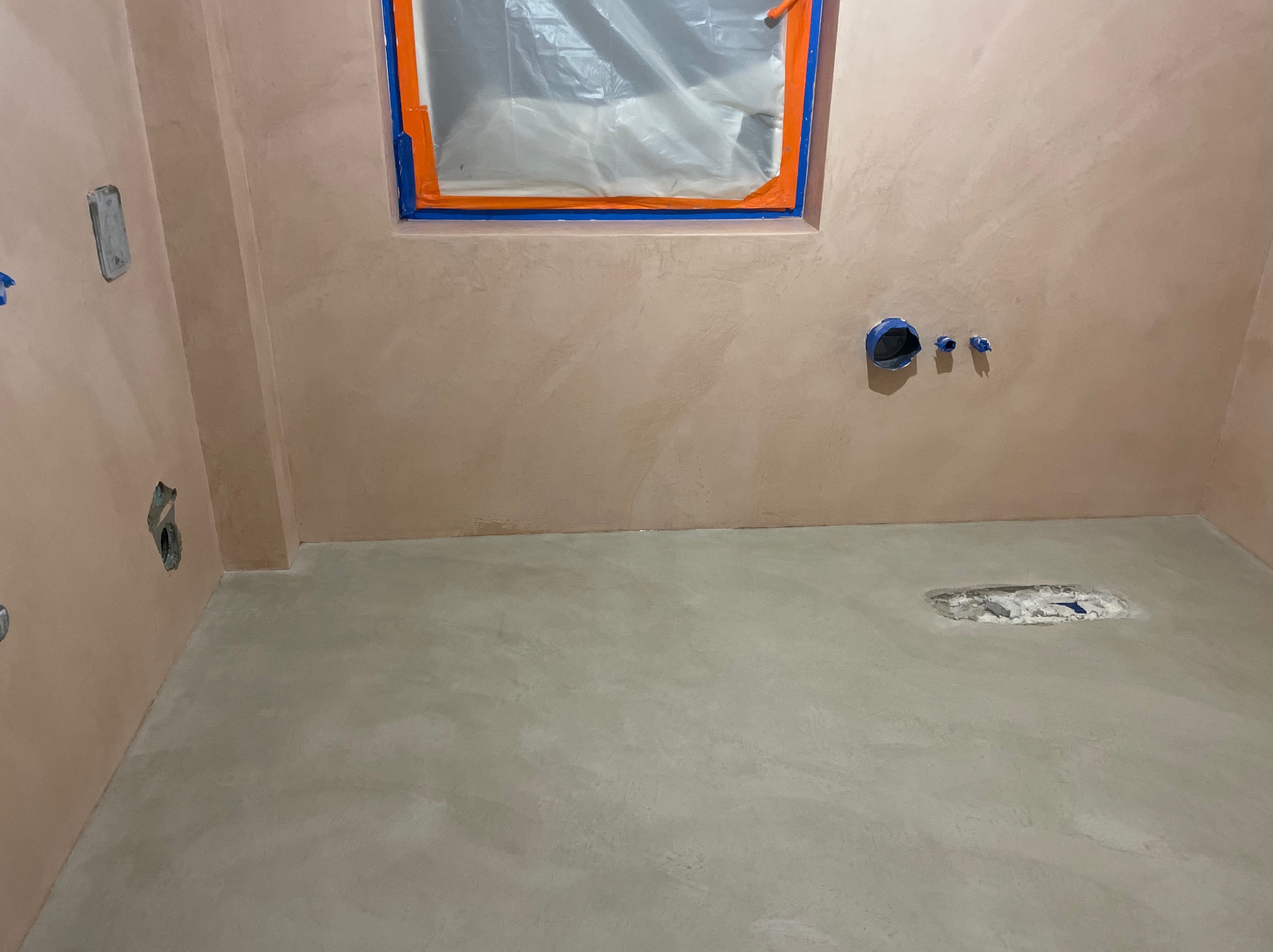 Przygotowanie łazienki do wykończenia, ściany pokryte beżowym tynkiem dekoracyjnym, zabezpieczone okno folią malarską, w podłodze otwór instalacyjny.