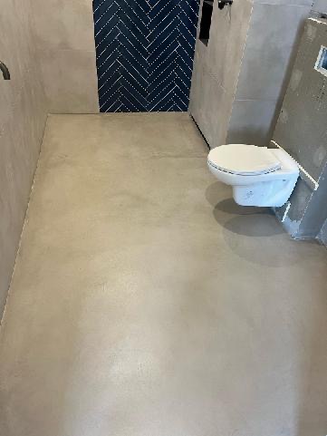 Wykończona łazienka z podłogą z mikrocementu w odcieniach beżu, kontrastująca z ciemnoniebieską ścianą w jodełkę i wiszącą toaletą.