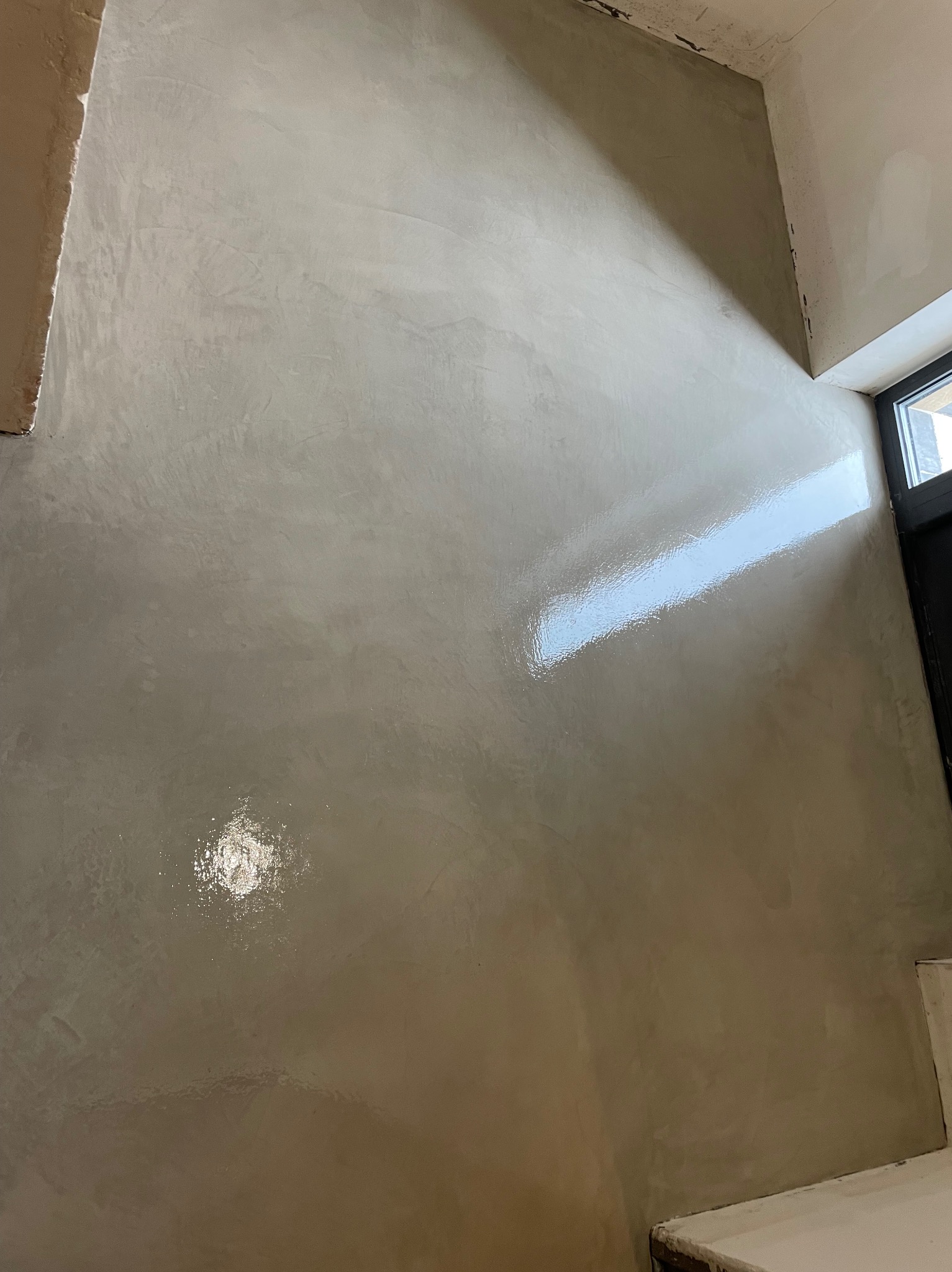 Ściana pokryta szarą, błyszczącą powłoką w minimalistycznym wnętrzu, z widocznymi refleksami światła i surowym wykończeniem narożników.