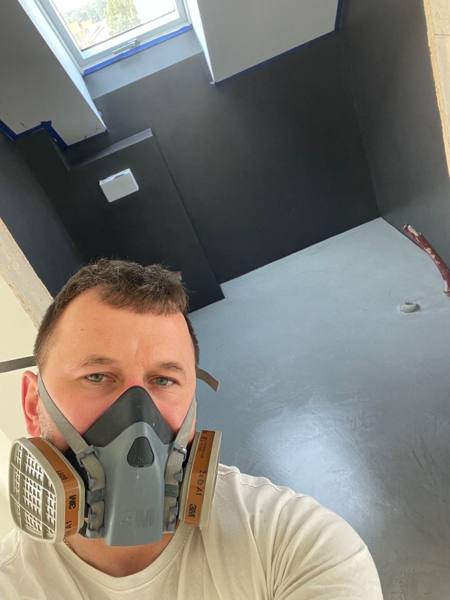 Pracownik w masce ochronnej na tle ciemnej, gładkiej powierzchni sufitu z oknem dachowym, zabezpieczonego taśmą malarską.