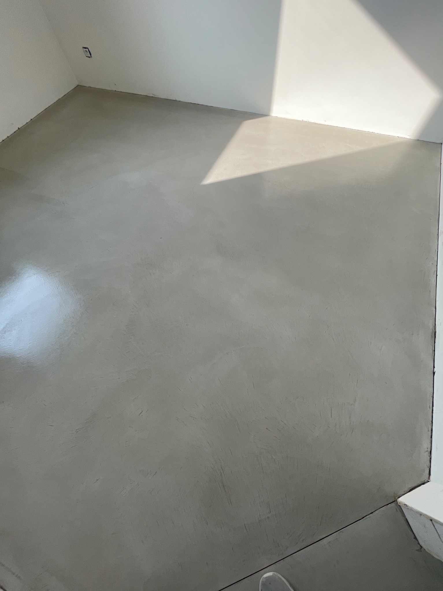 Jasnoszara, gładka posadzka w rogu pomieszczenia, oświetlona naturalnym światłem wpadającym przez okno, widoczne delikatne smugi i faktura na powierzchni.