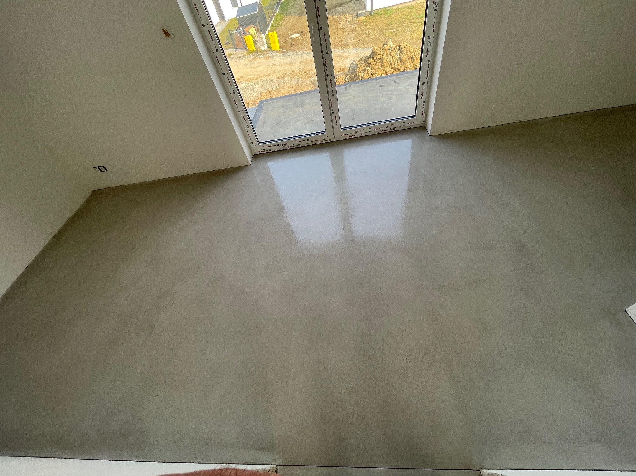 Gładka, szara posadzka w nowym pomieszczeniu z dużym oknem tarasowym, widok na plac budowy na zewnątrz.