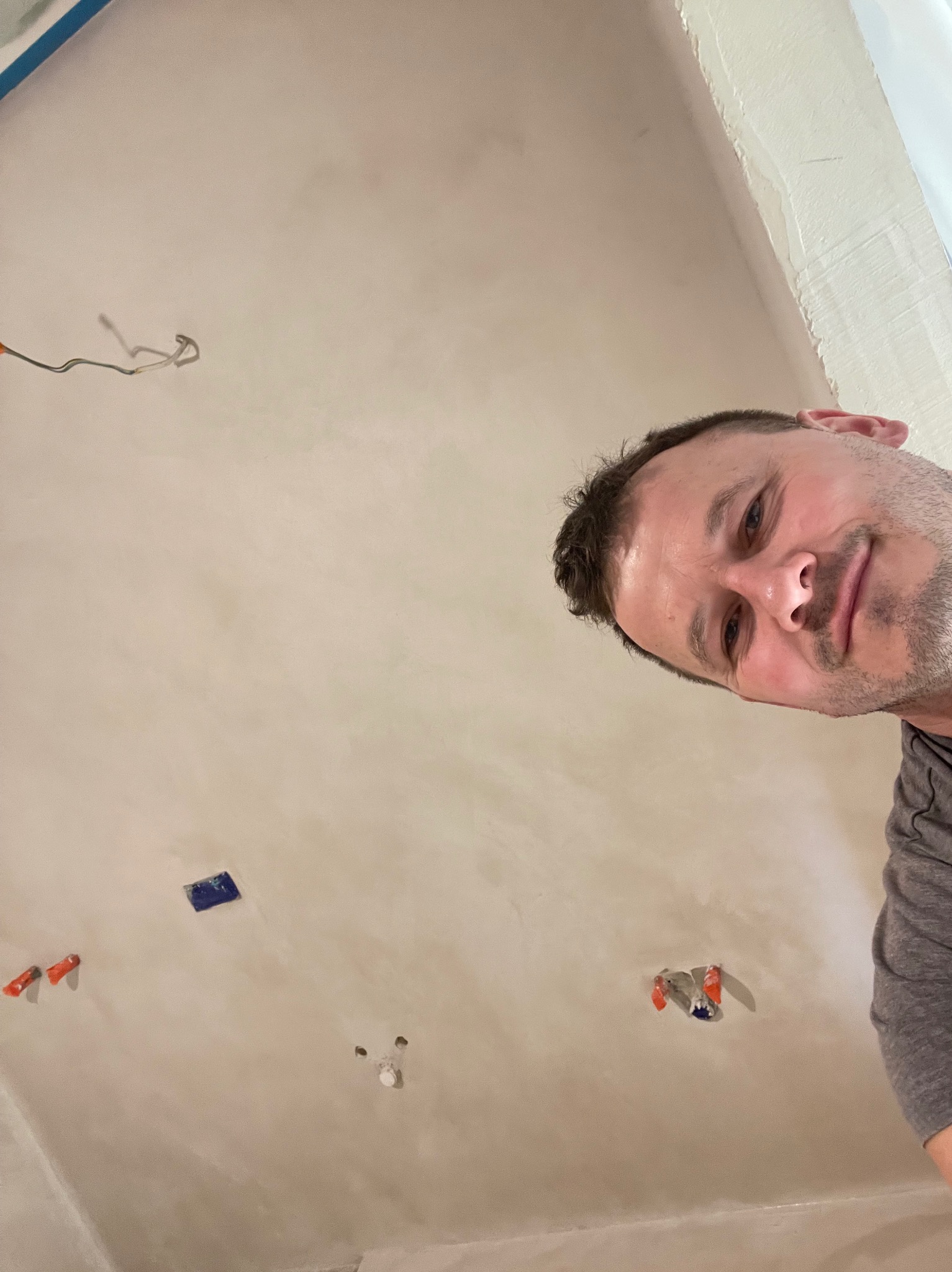 Widok z dołu na sufit w trakcie prac wykończeniowych, widoczne wystające przewody, zabezpieczone gniazdka i uśmiechnięta twarz wykonawcy.