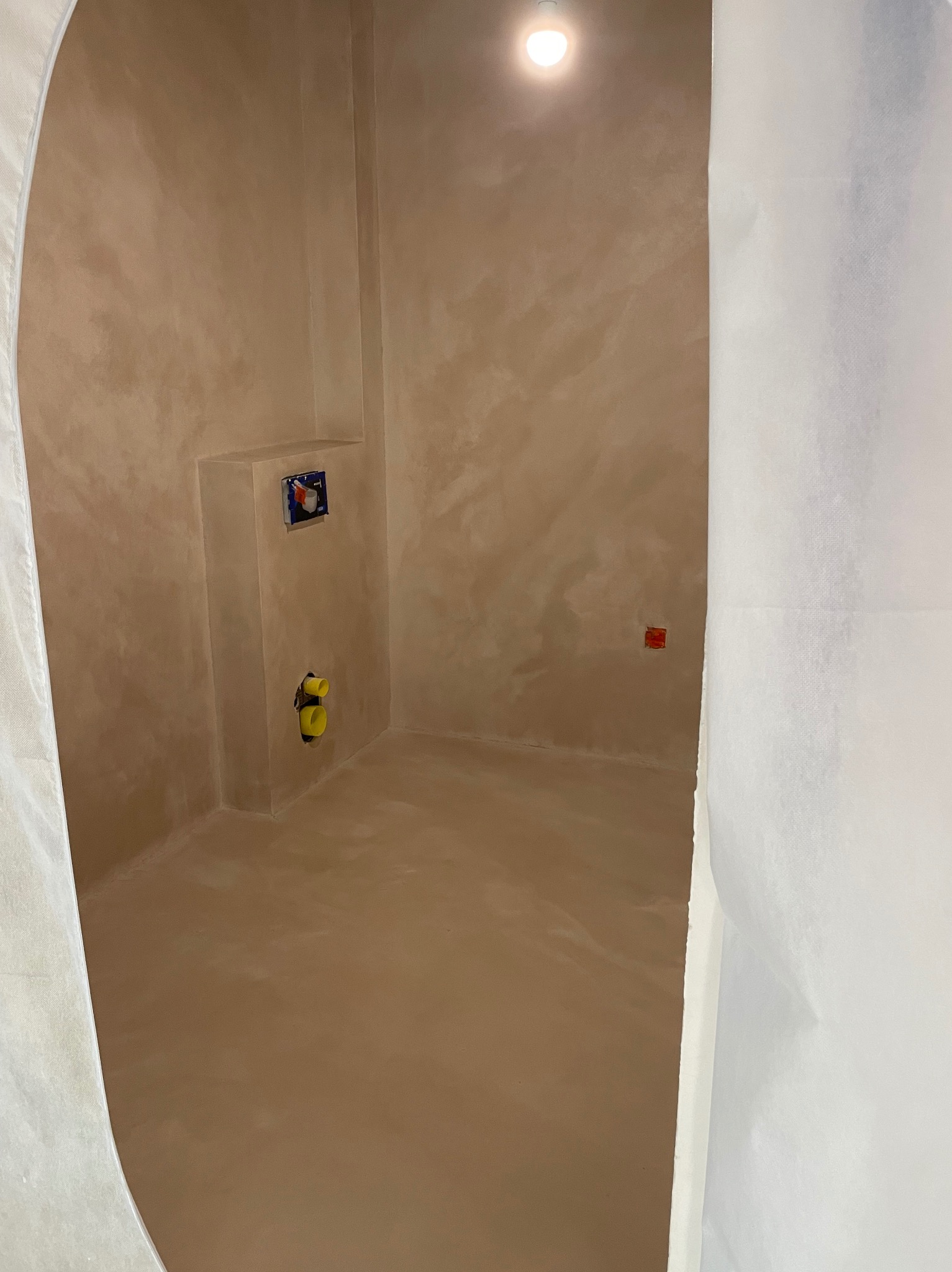 Niewielkie pomieszczenie wykończone beżową, teksturowaną powłoką; widoczne instalacje wodno-kanalizacyjne i elektryczne, osłonięte folią malarską.