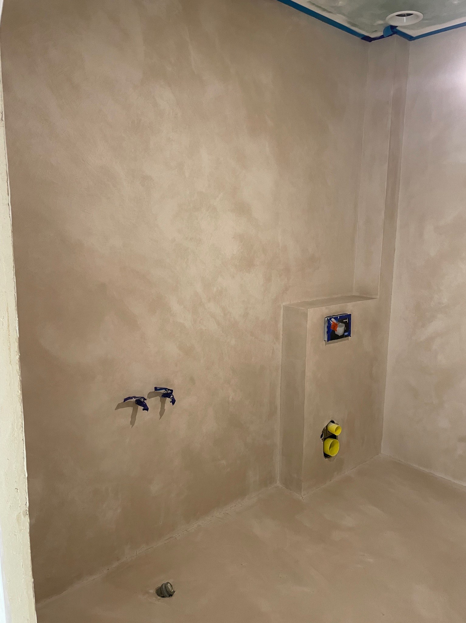 Beżowa ściana i podłoga w łazience wykończone z efektem dekoracyjnym, widoczne instalacje wodno-kanalizacyjne i niebieska taśma malarska przy suficie.