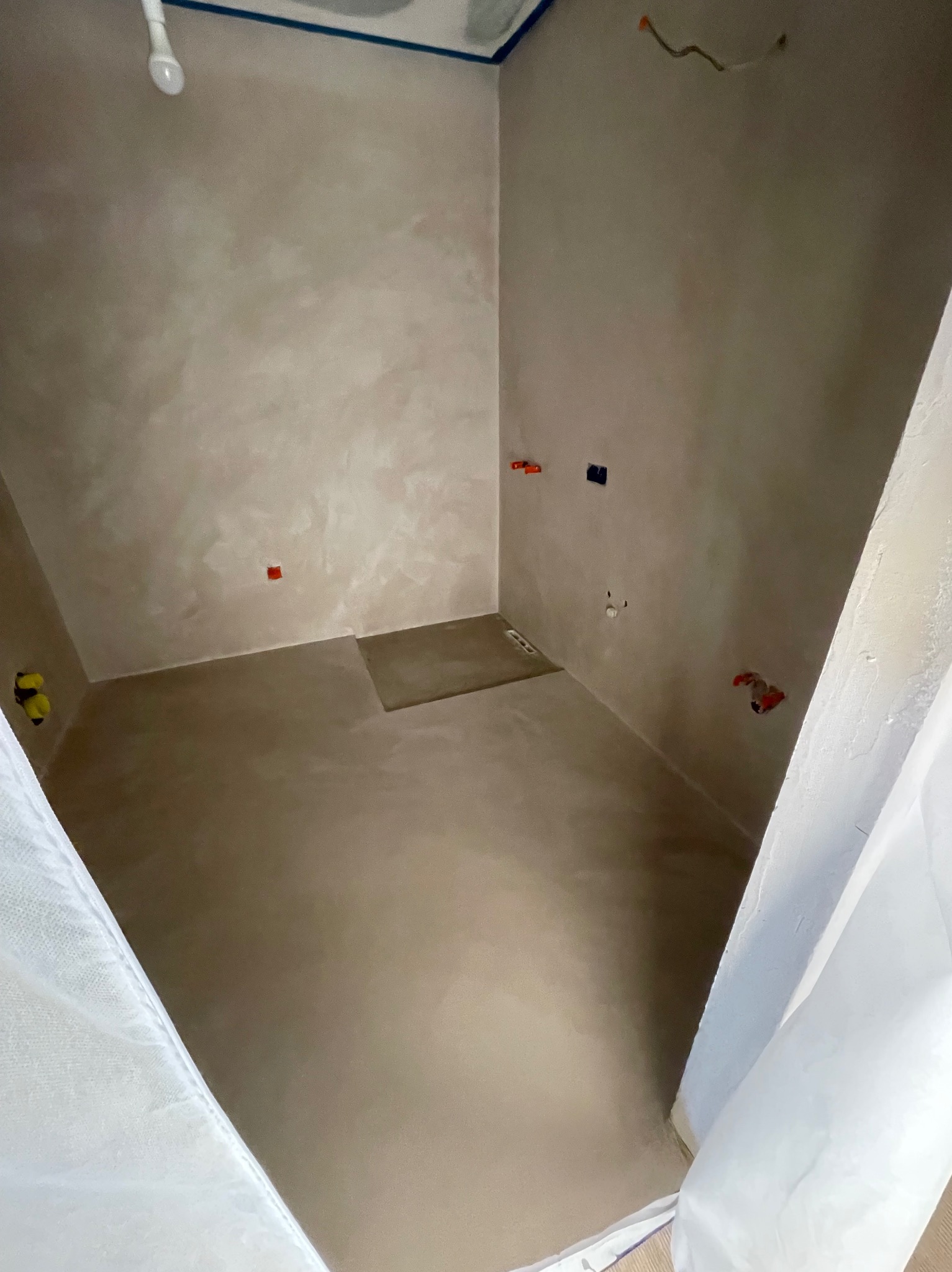 Wnętrze łazienki w trakcie wykańczania, ściany i podłoga pokryte beżową, gładką powierzchnią, widoczne instalacje elektryczne i wodne, zabezpieczone folią fragmenty pomieszczenia.