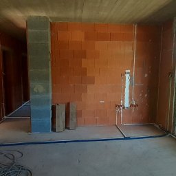 EL- BAR Bartosz Ostrowski - Surowy stan wnętrza z instalacjami elektrycznymi i hydraulicznymi w trakcie montażu, widoczne kable i rury na ścianach z czerwonej cegły, betonowy sufit i okno z widokiem na zieleń.