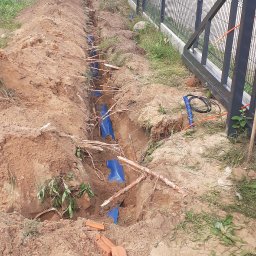 EL- BAR Bartosz Ostrowski - Wykop w ziemi z ułożonymi niebieskimi rurami osłonowymi, widoczne fragmenty cegieł i korzenie drzew, w tle metalowe ogrodzenie panelowe i zieleń.