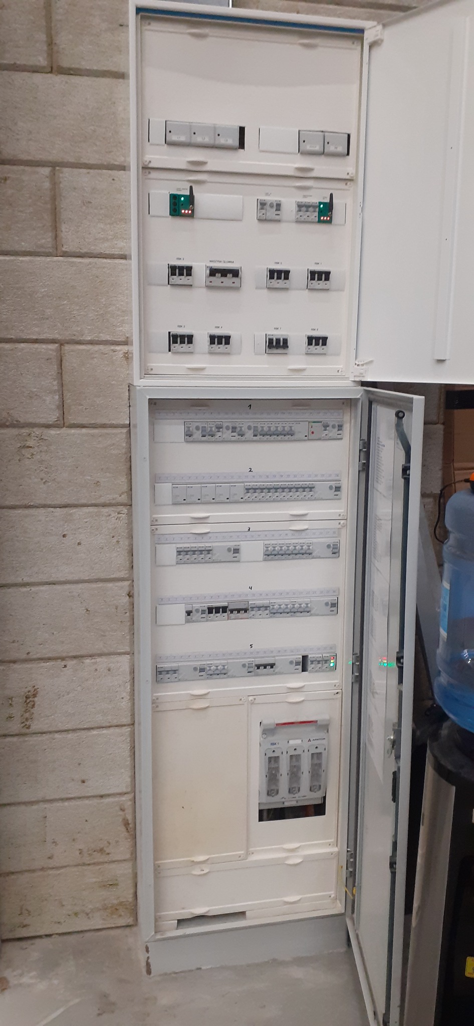 Otwarta skrzynka elektryczna z wieloma bezpiecznikami i modułami, umieszczona na ścianie z bloków betonowych, widoczna butla z wodą w tle.