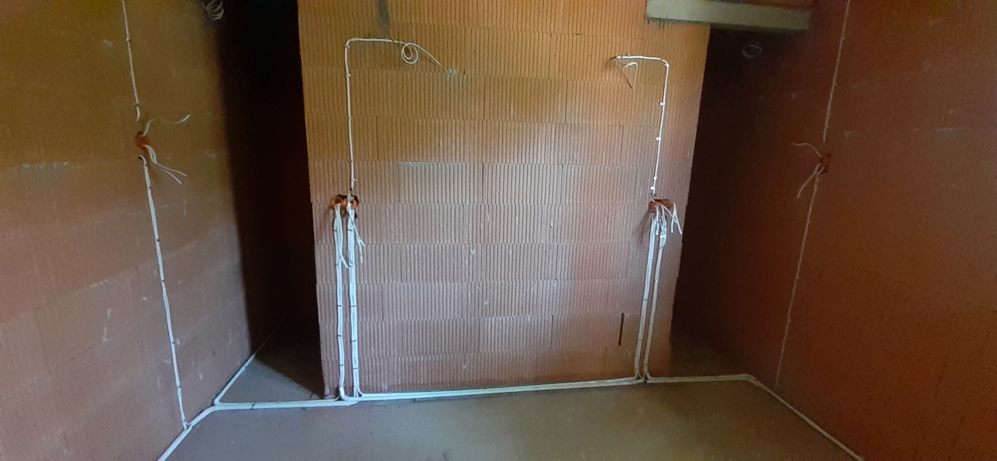 Surowy mur z instalacją elektryczną w trakcie montażu, widoczne kable wystające ze ścian i prowadzone wzdłuż podłogi, brak wykończenia wnętrza.