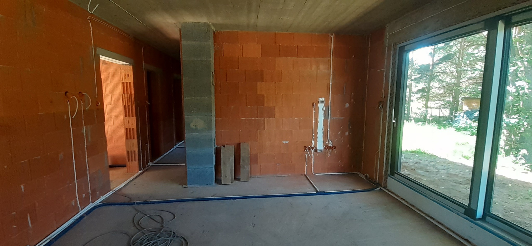 Surowy stan wnętrza z instalacjami elektrycznymi i hydraulicznymi w trakcie montażu, widoczne kable i rury na ścianach z czerwonej cegły, betonowy sufit i okno z widokiem na zieleń.