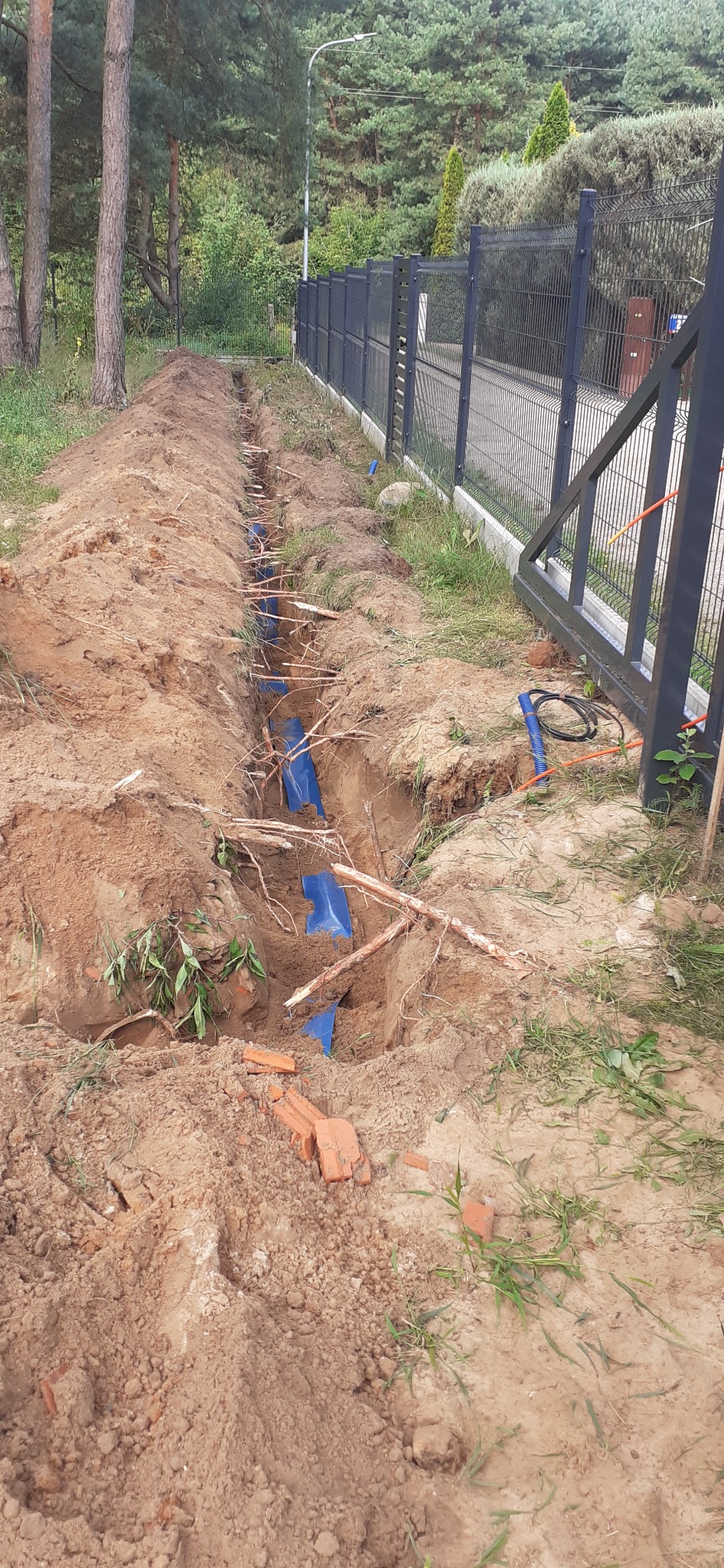 Wykop w ziemi z ułożonymi niebieskimi rurami osłonowymi, widoczne fragmenty cegieł i korzenie drzew, w tle metalowe ogrodzenie panelowe i zieleń.