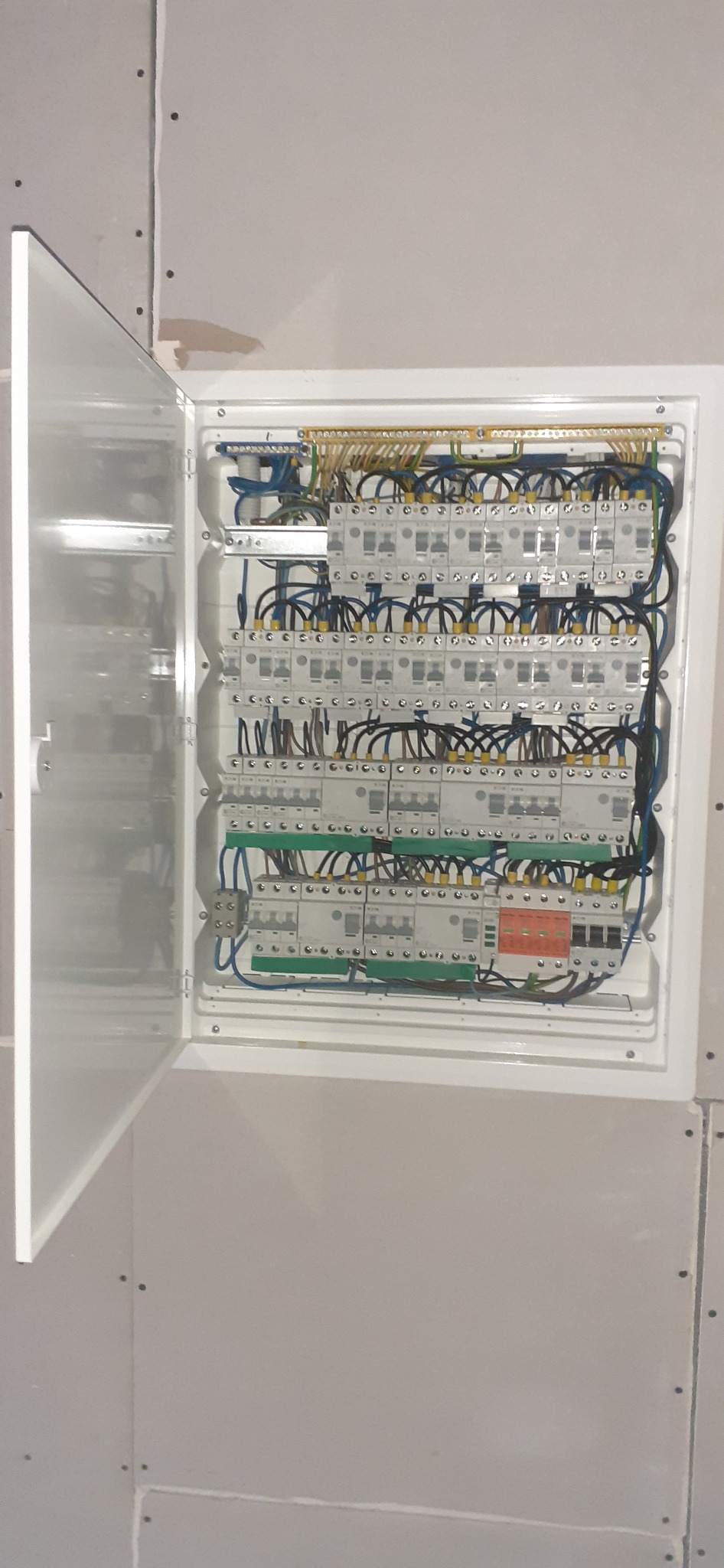 Otwarta skrzynka elektryczna z bezpiecznikami i okablowaniem zamontowana na ścianie pokrytej płytami gipsowo-kartonowymi.