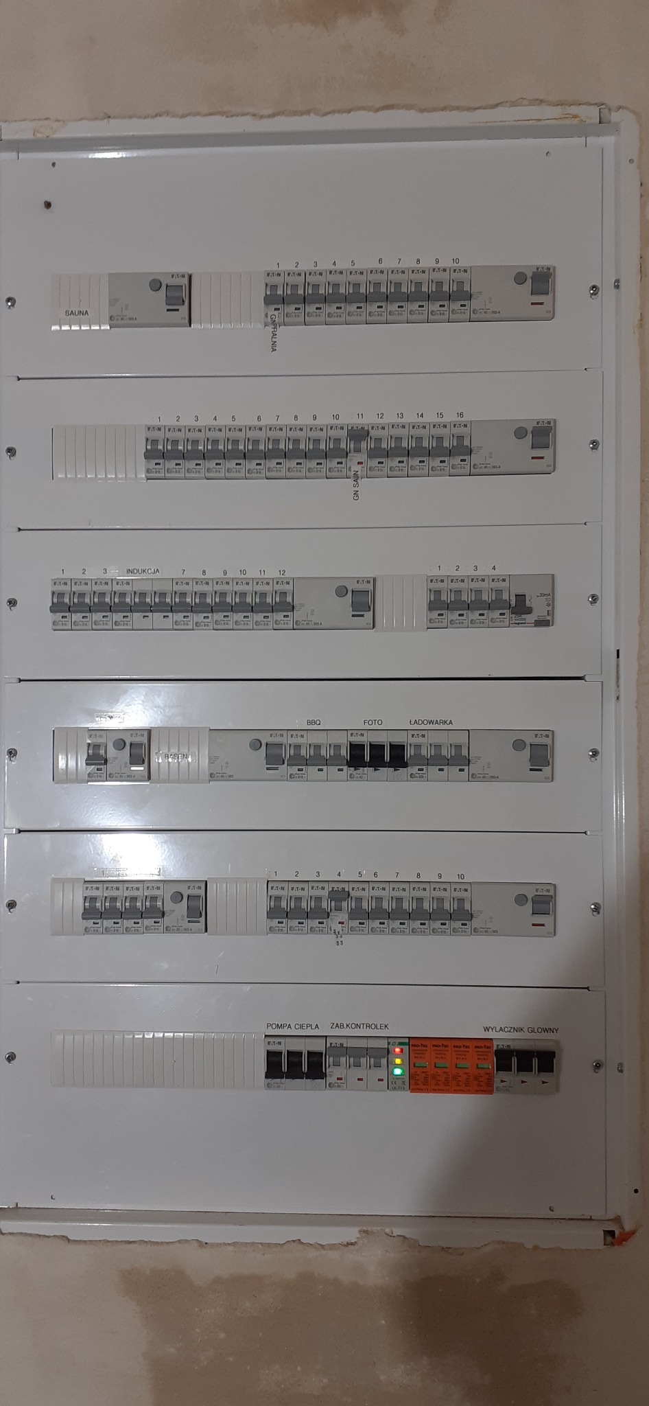 Rozdzielnica elektryczna z opisami obwodów (sauna, indukcja, BBQ, foto, ładowarka, pompa ciepła, zab.kontrolek, wyłącznik główny) i zainstalowanymi bezpiecznikami automatycznymi, widok z bliska...