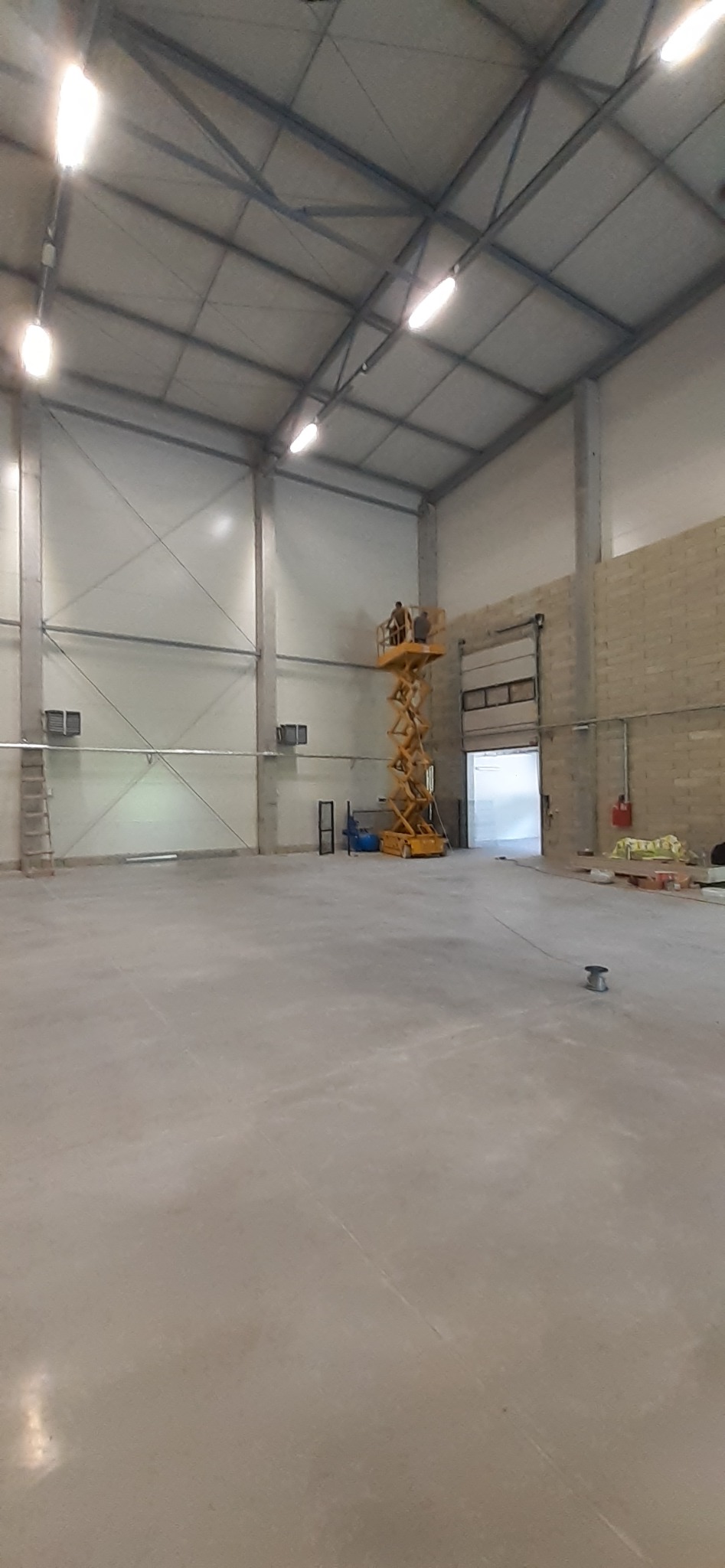 Dwóch pracowników na podnośniku nożycowym montuje oświetlenie w hali magazynowej z betonową podłogą i metalową konstrukcją dachu.