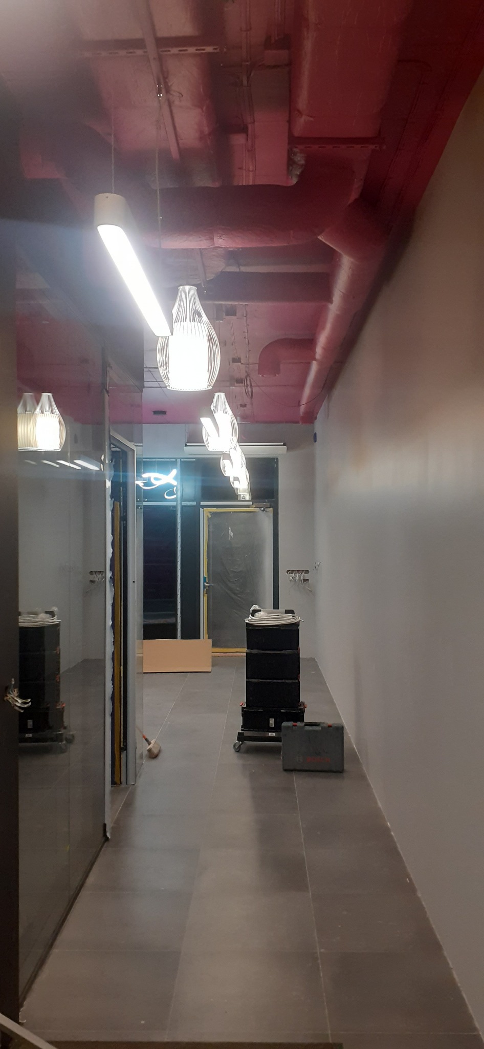 Wnętrze korytarza w trakcie remontu z widocznymi lampami wiszącymi, odkrytymi rurami pod sufitem, skrzynkami narzędziowymi i śladami po instalacjach elektrycznych na ścianie.
