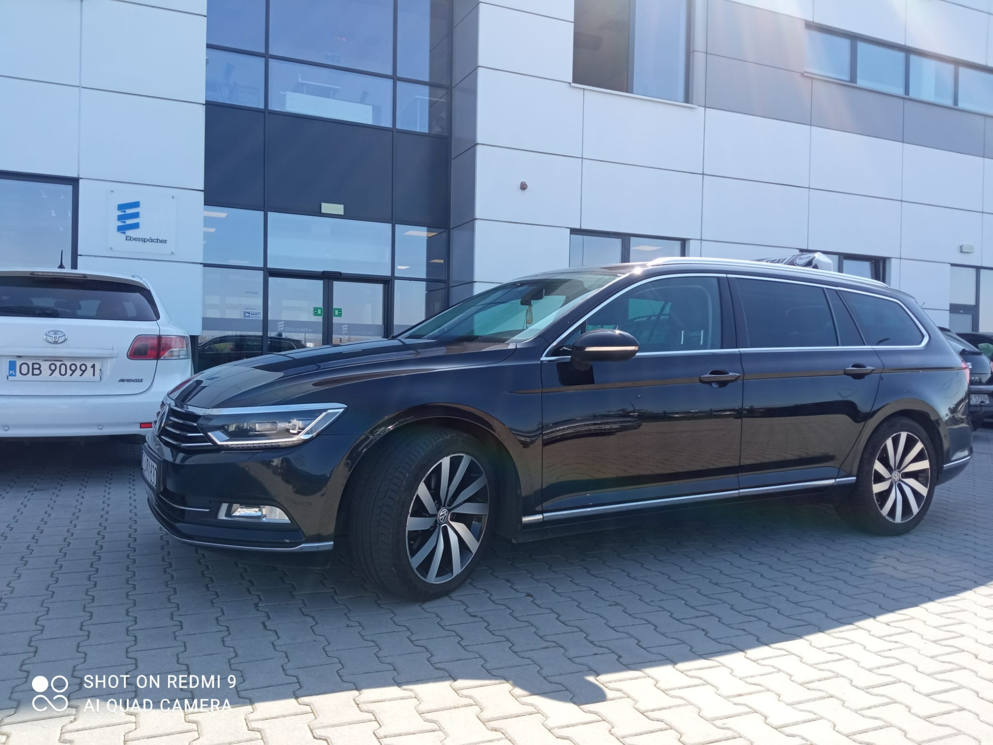 Czarny samochód kombi marki Volkswagen Passat zaparkowany na kostce brukowej przed nowoczesnym budynkiem biurowym z dużymi oknami.