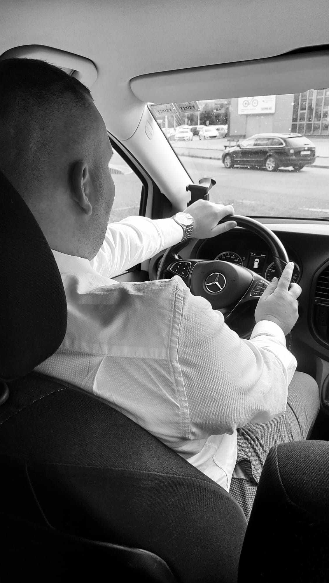 Kierowca w białej koszuli prowadzi Mercedesa Vito, widoczny zegarek na ręce i ulica za szybą.
