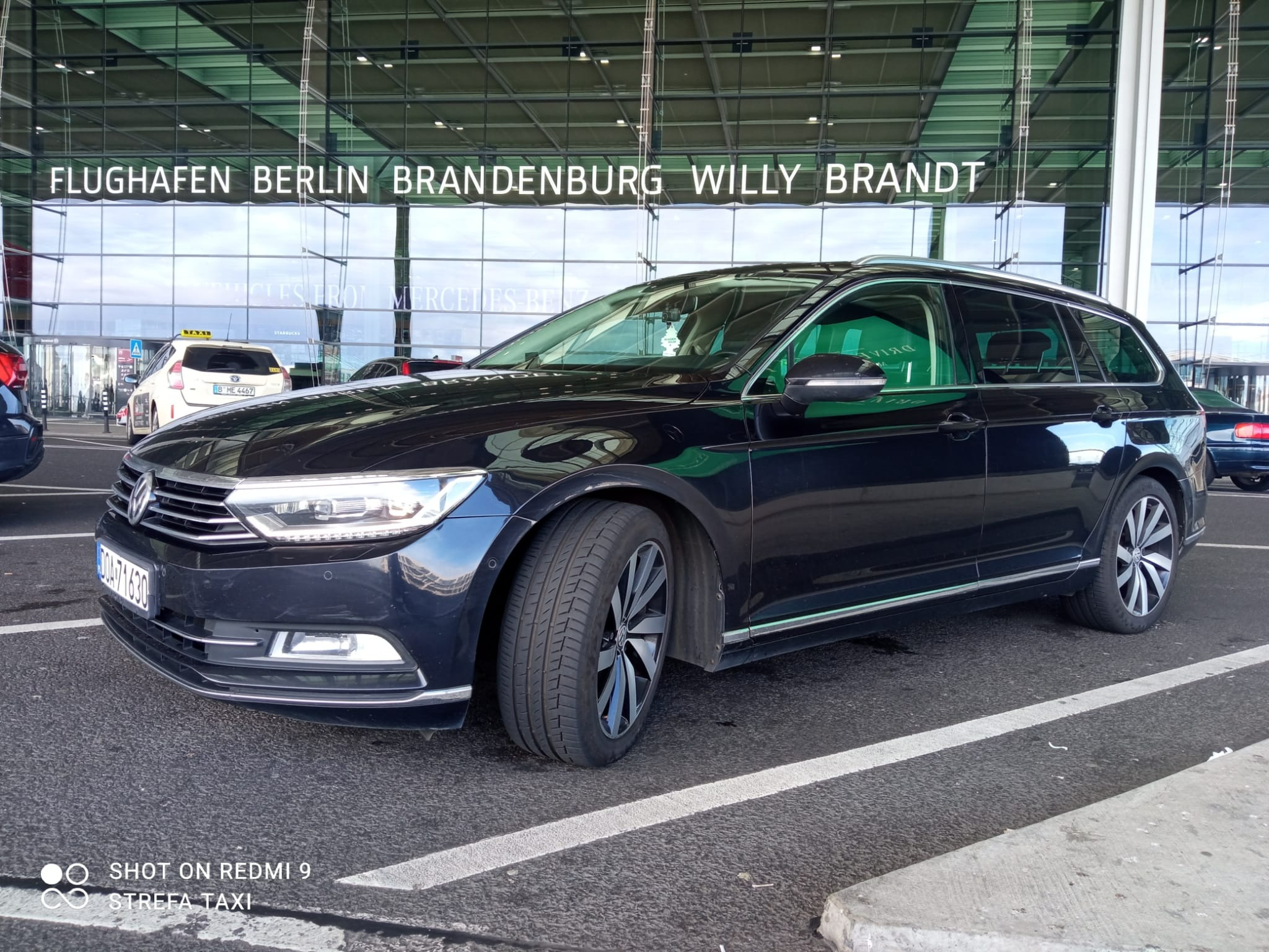 Czarny samochód kombi marki Volkswagen Passat stoi przed szklanym budynkiem lotniska Berlin Brandenburg Willy Brandt, w tle widoczna taksówka.