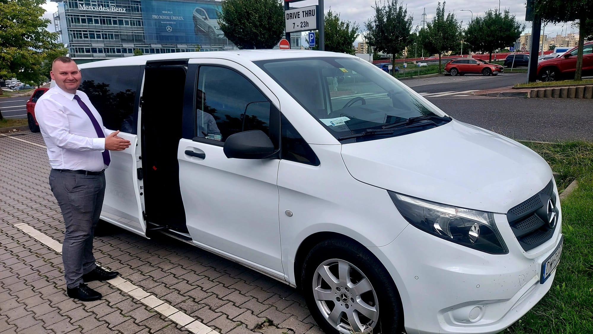 Kierowca w koszuli i krawacie gestykuluje przy otwartych drzwiach białego busa Mercedes Vito, zaparkowanego na kostce brukowej. W tle budynek Mercedes-Benz.