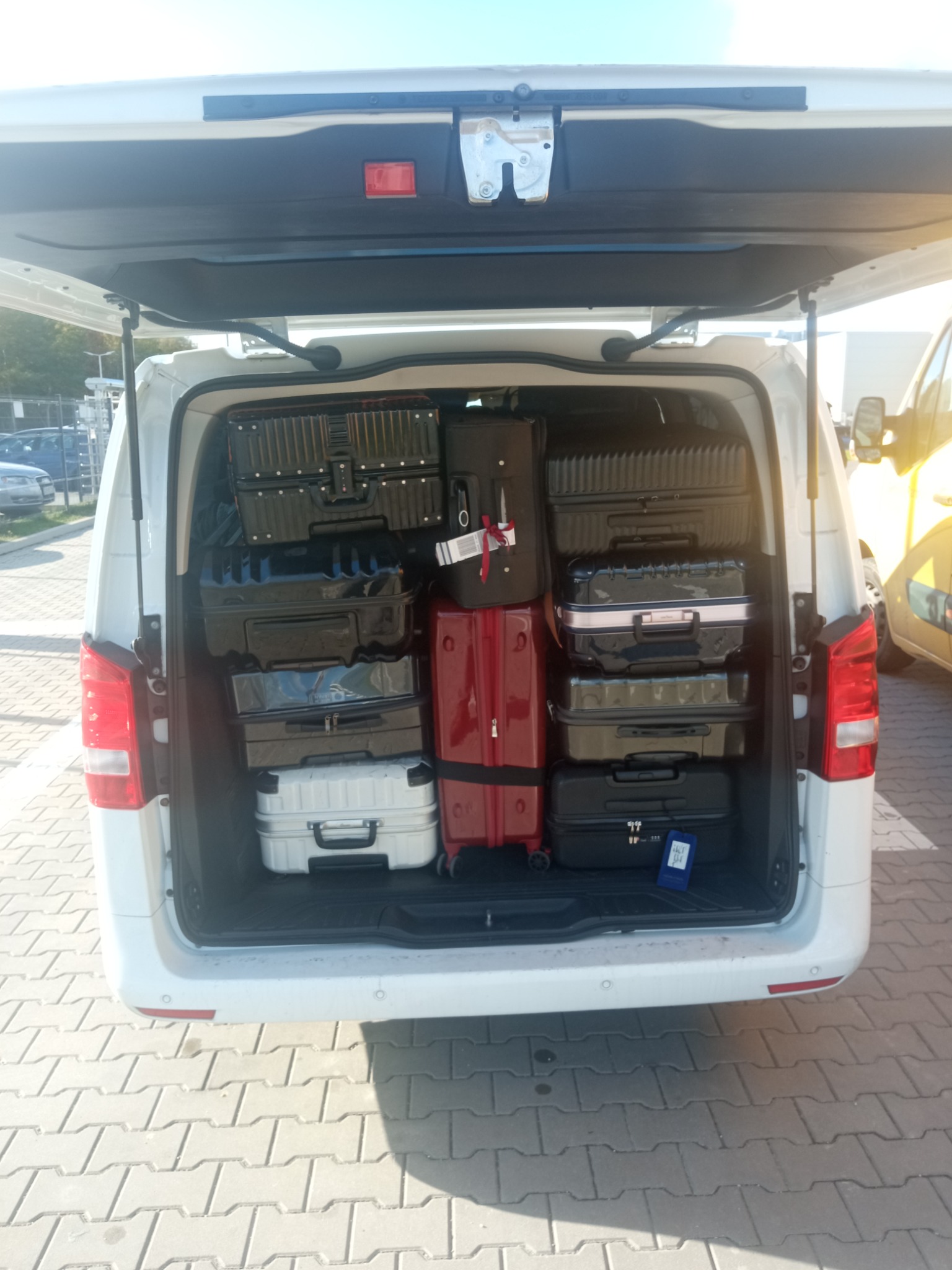 Bagażnik białego vana wypełniony po brzegi różnymi walizkami, w tym czarnymi, czerwonymi i srebrnymi, z otwartą klapą bagażnika.